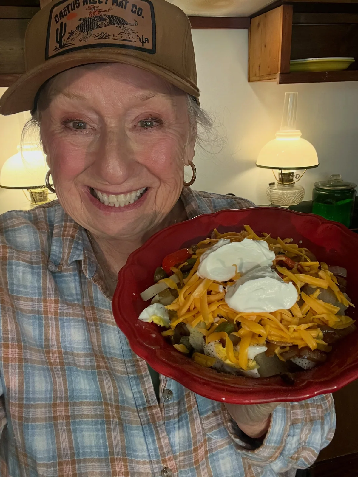 A smiling home cook holding a red bowl of potatoes with shredded cheese, colorful toppings, and a creamy white topping