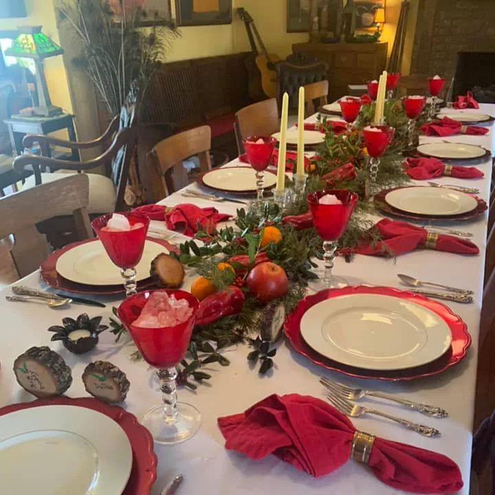 A long holiday table set with white plates, red glasses, candles, greenery and fruit
