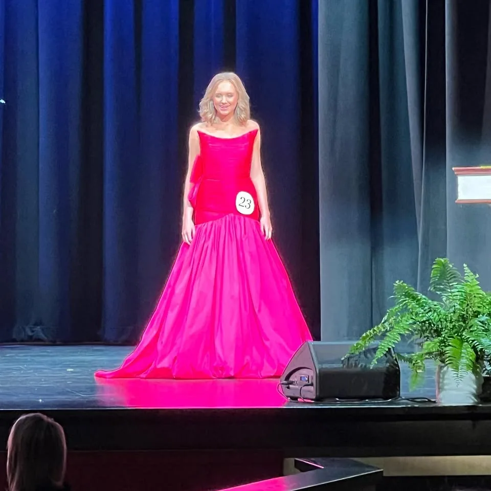 A young woman in a vivid pink formal gown walking across an auditorium stage